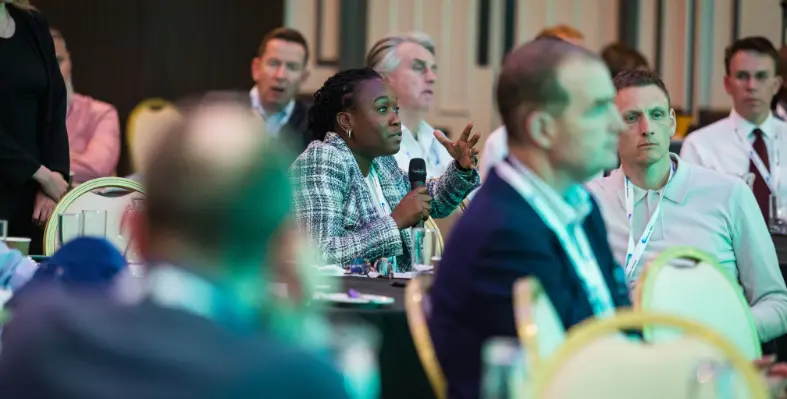 A lady with a microphone mid-conversation at a conference with other delegates sat around her